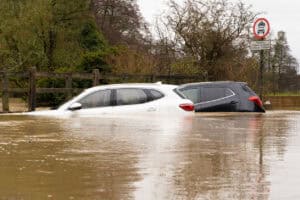 Two cars submerged in muddy flood water on a flooded UK road, showing the kind of flooding a flood risk assessment is required to address.