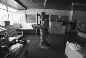 A man cleaning up after a flood wades through floodwater in an office, rescuing files and paperwork from the rising water.