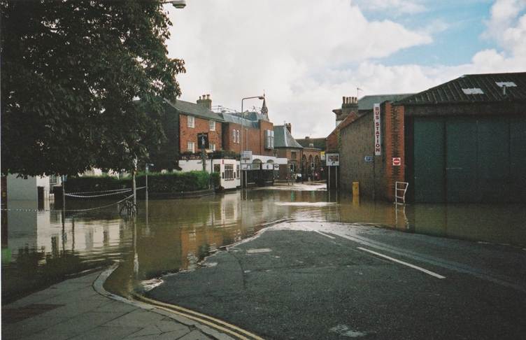 A flooded street in a town with water covering the road and reaching the entrances of brick buildings on both sides; cloudy sky above and reflections in the water.