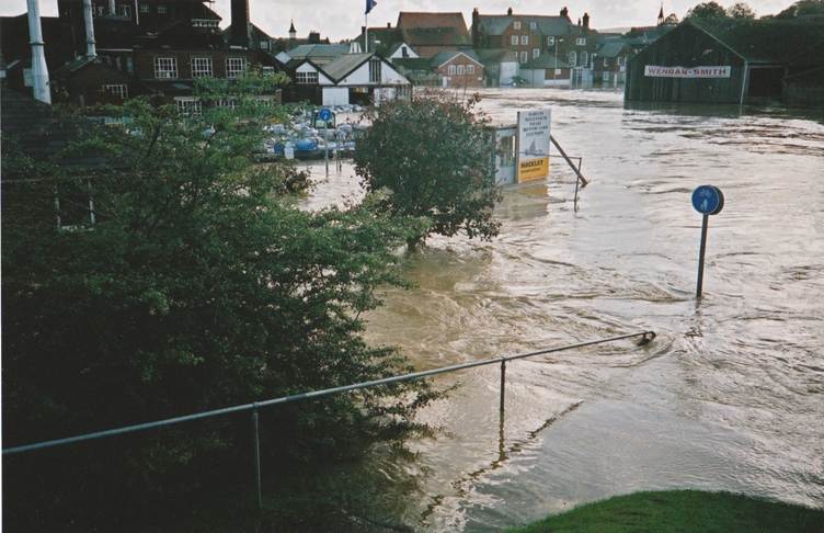 Flooded street in a residential area with brown water submerging road signs, railings, and parts of buildings. Trees and houses are partially surrounded by floodwater under a cloudy sky.