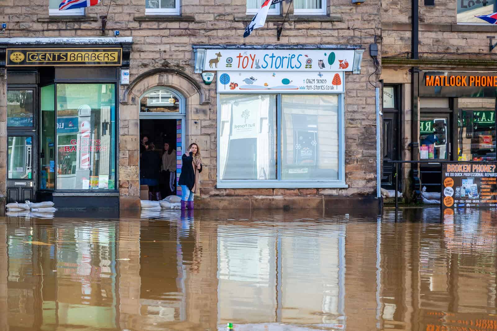 Flooded UK high street with sandbags at shop doorways and a resident standing in wellies outside a small business.