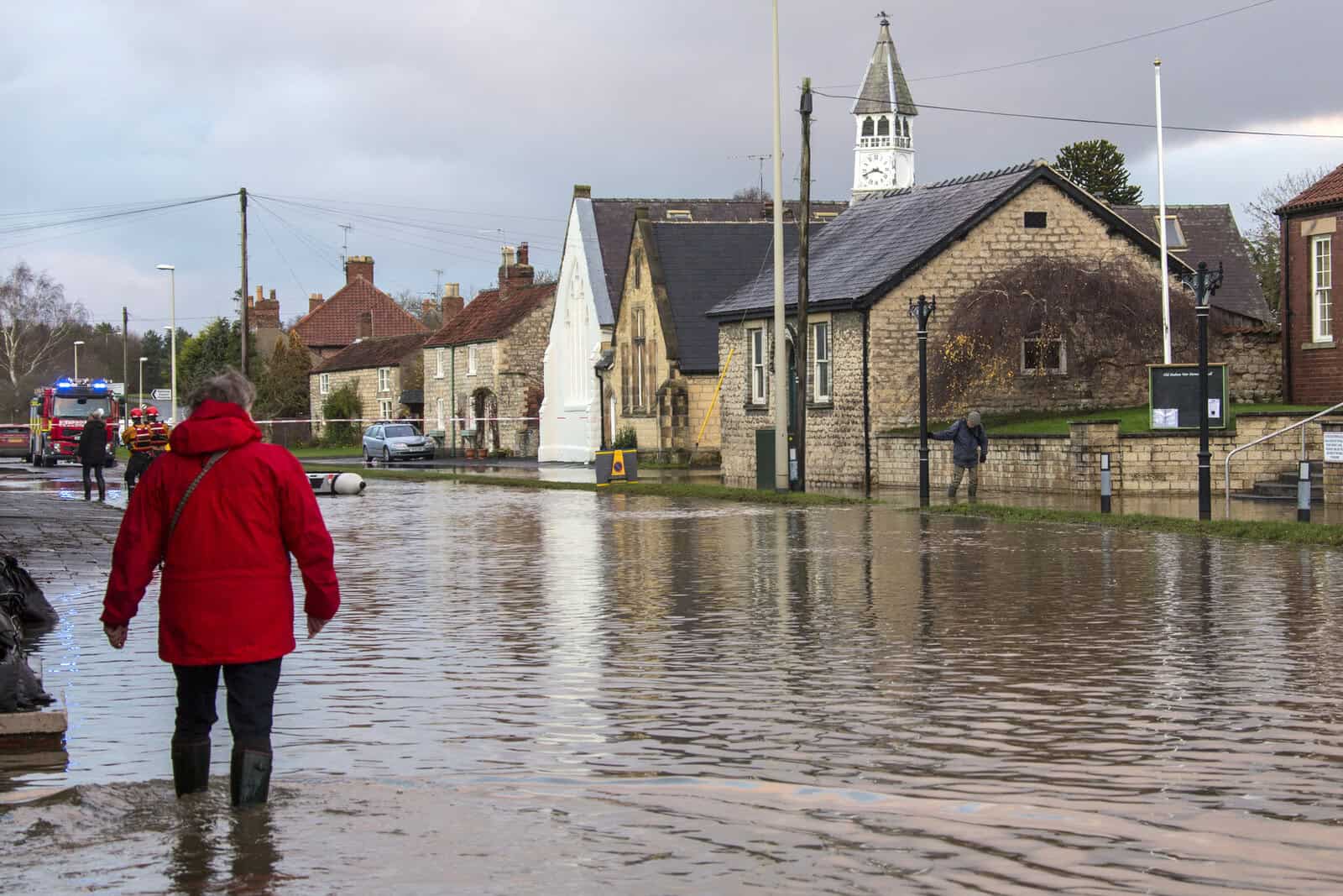 Resident wading through floodwater on a UK village street with emergency services and stone buildings behind.