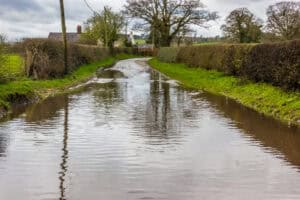 Standing floodwater across a rural lane between hedgerows, showing the prolonged flooding that saturated ground can cause, demonstrating the need for a groundwater flood risk assessment.