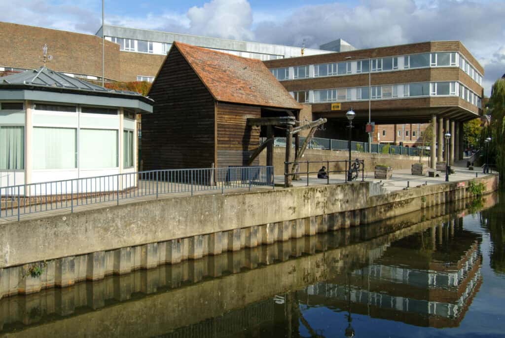 View along the River Wey in central Guildford, with a historic timber warehouse and crane on the quayside beside modern town-centre buildings.