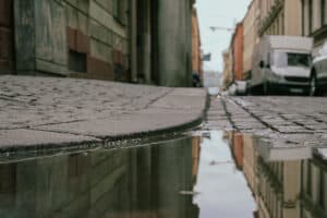 Surface water pooled on a cobbled urban street, reflecting nearby buildings after rainfall.