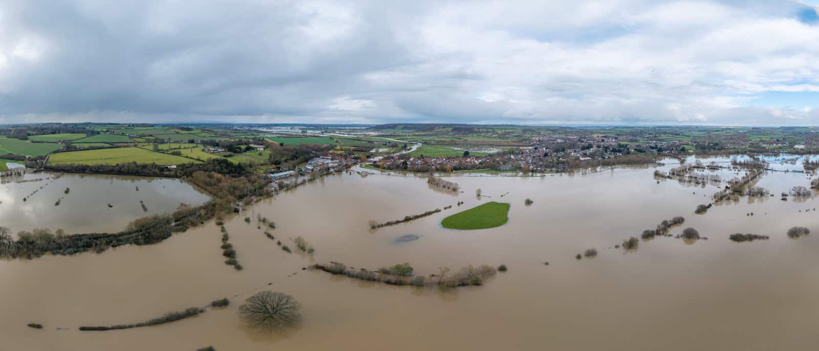 Aerial view of a flooded UK village showing brown floodwater spread across surrounding farmland, with houses on slightly higher ground.