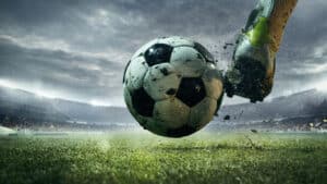 Boot striking a mud-flecked ball on a floodlit stadium pitch, the wet playing surface that football pitch drainage keeps firm.