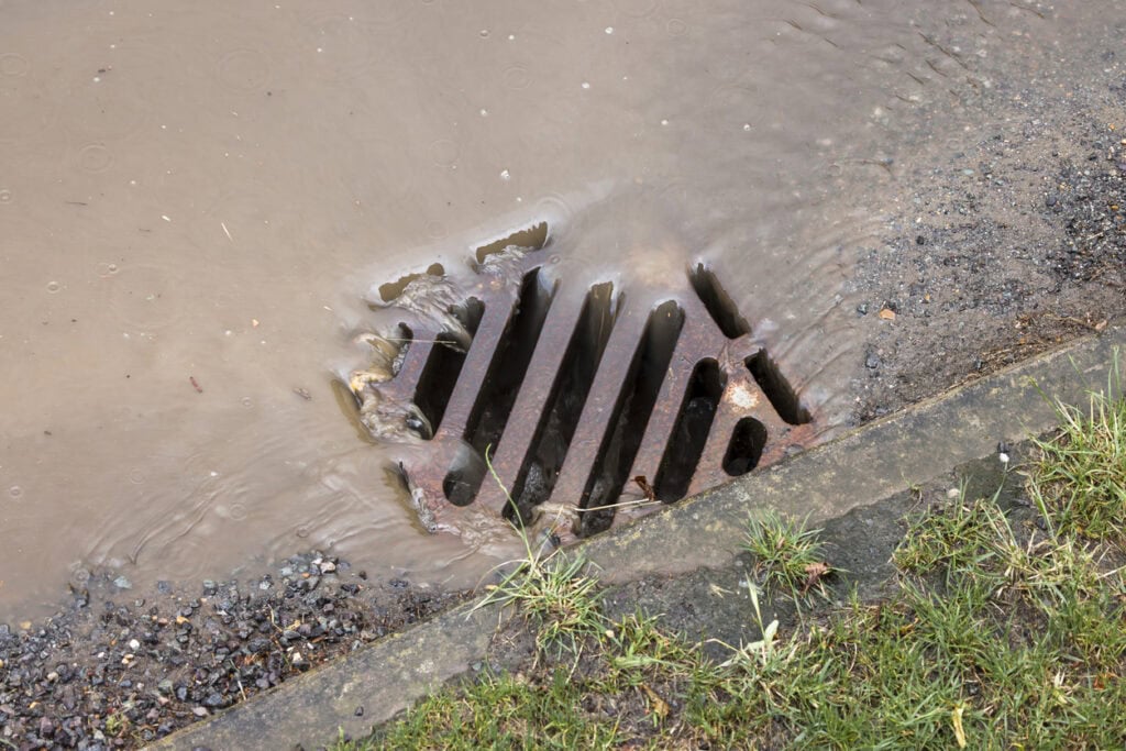 Muddy first flush run-off draining off a road into a kerbside gully grate during heavy rain.