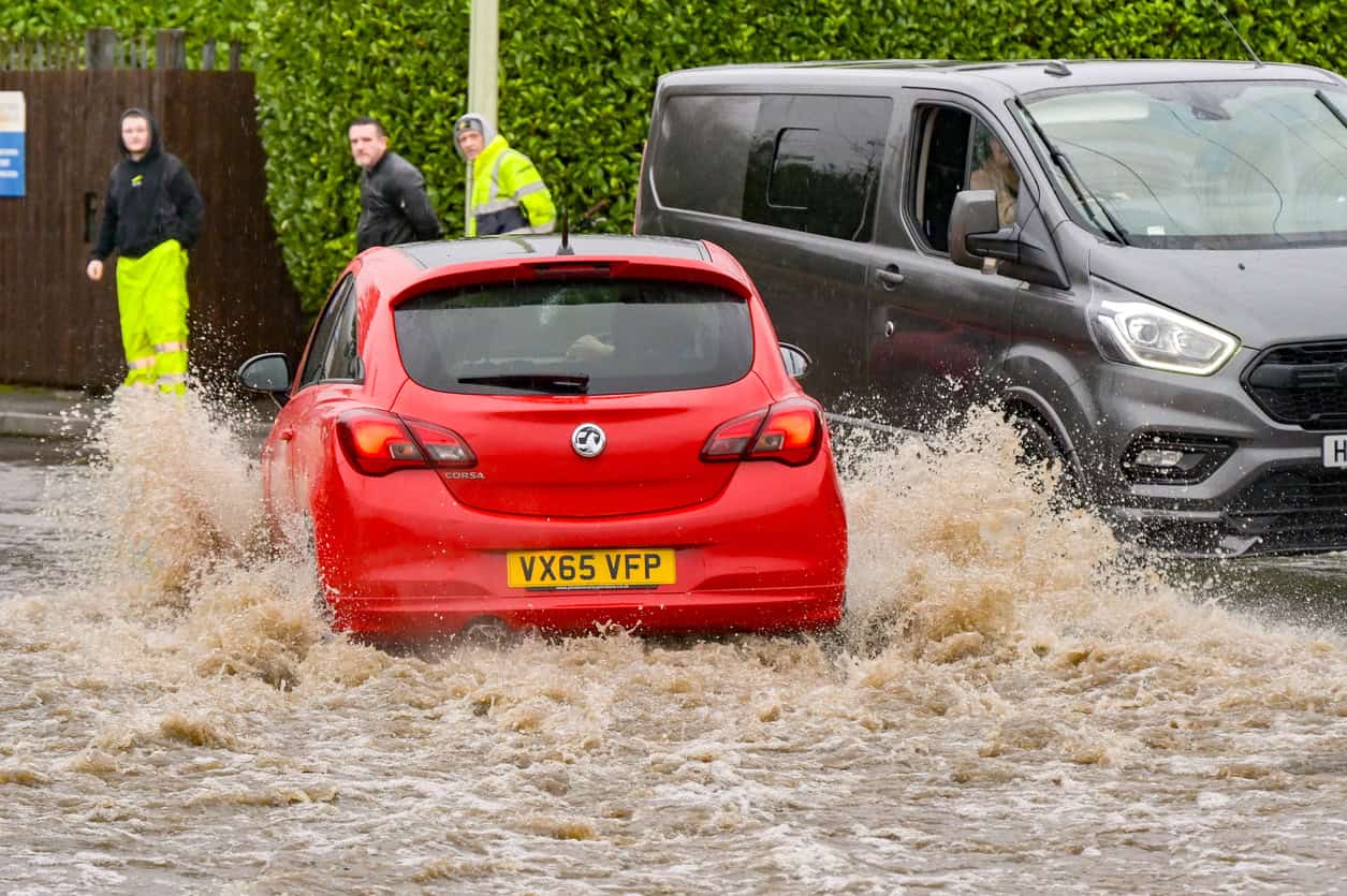 A red hatchback driven fast through brown urban flood water, throwing a heavy spray towards pedestrians standing on the pavement.