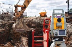 Excavator digging a drainage trench by stacked pipes and a survey instrument, the groundworks a Section 106 sewer connection needs.