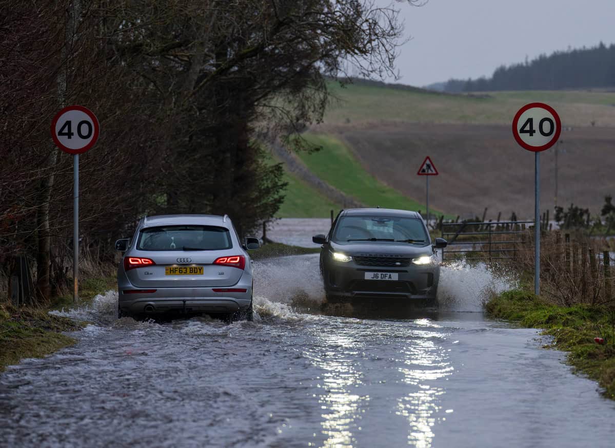 Two cars passing each other on a flooded rural road, one crossing slowly and the other throwing a wide spray across the carriageway.