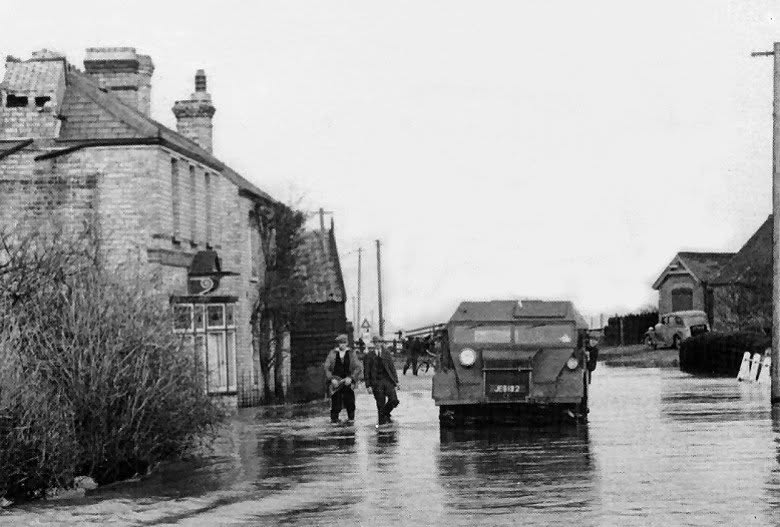 Men wading past a lorry in a flooded village street during the 1947 floods, beneath a chimney broken by the gale.