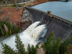 Concrete dam releasing water through its scour valves, the kind of raised structure breach modelling data is prepared for.