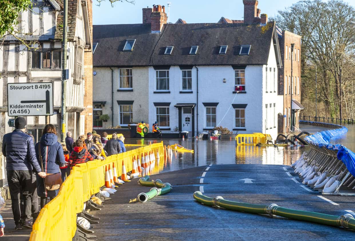 Temporary flood defences and pump hoses along a flooded street in Bewdley, with people watching from behind the barrier.