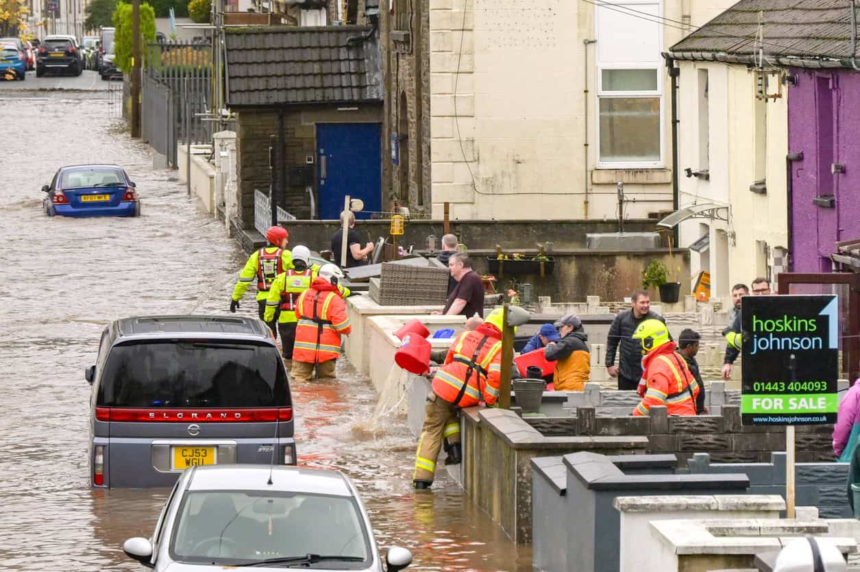 Rescue crews wading along a flooded street of older terraced houses, with cars standing in floodwater and a For Sale board outside one home