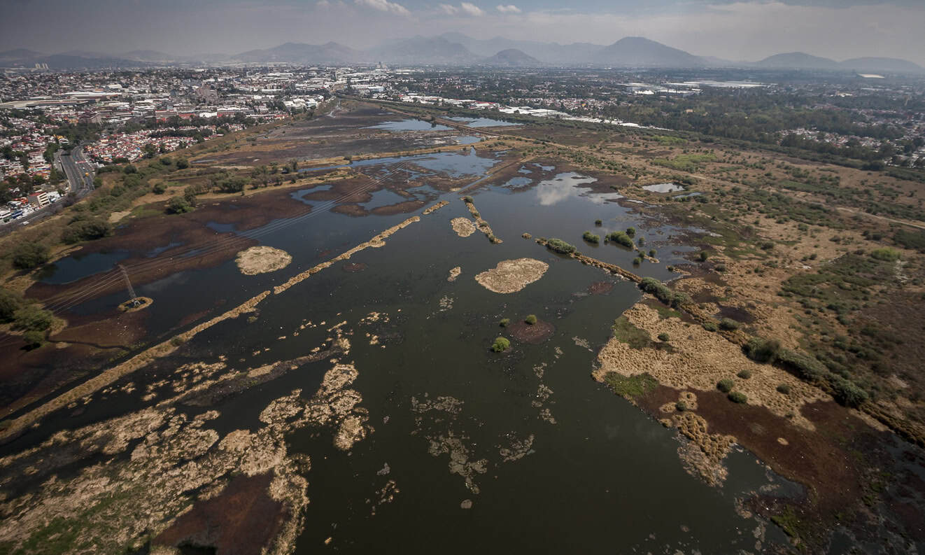 Shallow water and engineered bunds on the saline lakebed at the city's edge, the drained ground behind the Mexico City sinking.