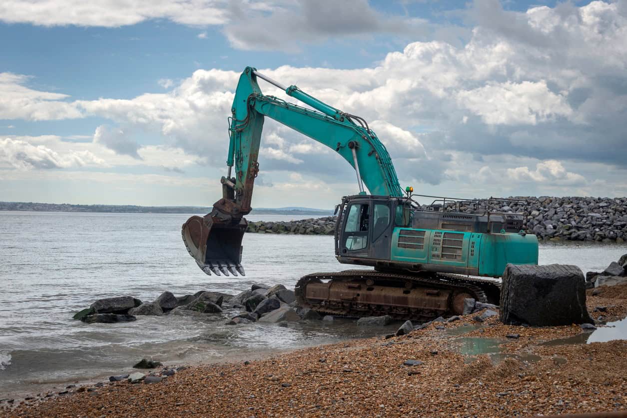Tracked excavator working on a rock breakwater on a shingle beach, a common hard defence against coastal erosion.