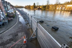 Demountable flood barriers braced along a riverside street, holding back a river in flood with seepage pooling behind them.